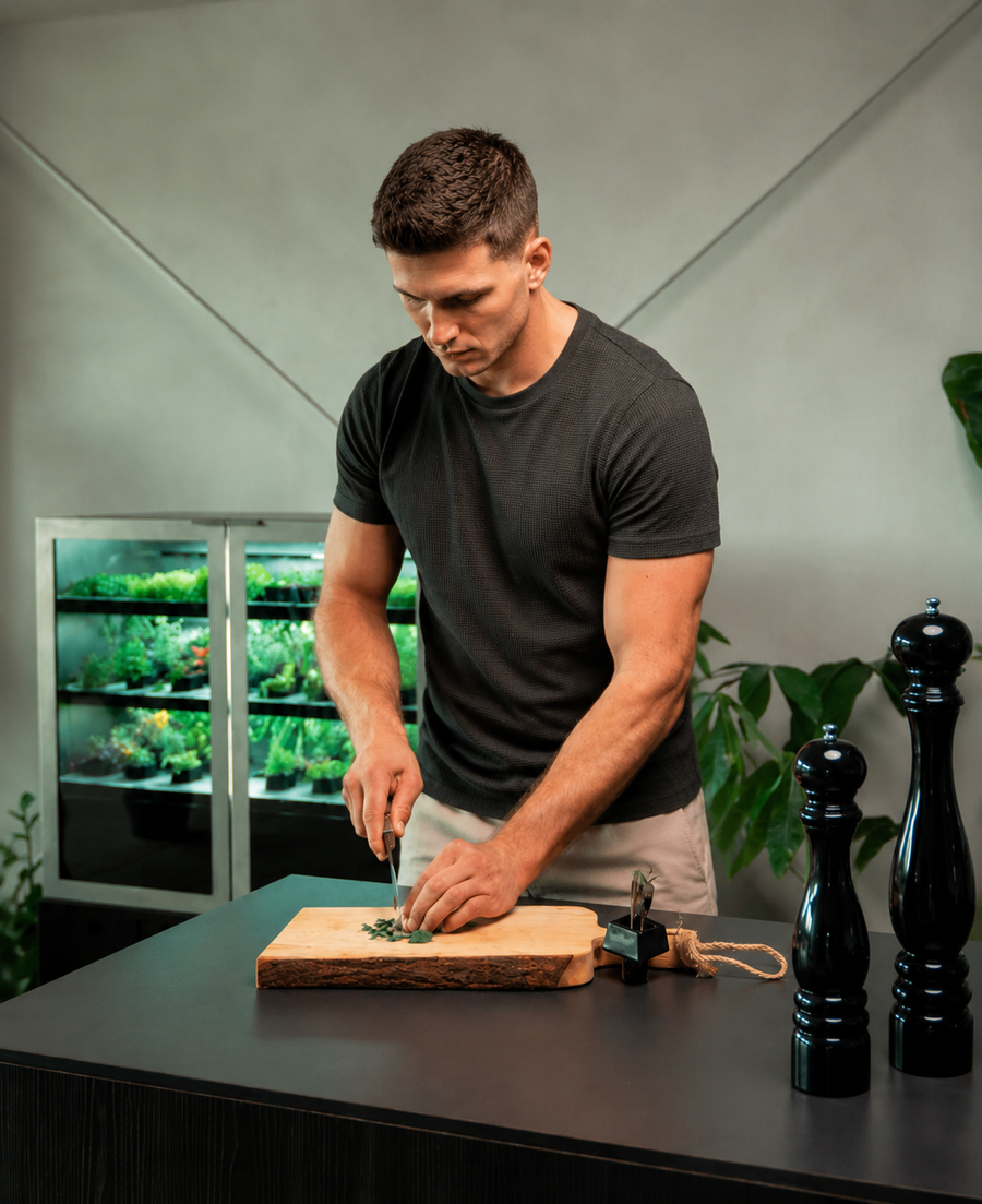 A focused young man in a dark textured T-shirt and light shorts chops fresh microgreens on a wooden cutting board atop a dark kitchen island. Behind him, illuminated shelves of growing greens sit inside a modern indoor gardening cabinet, while black pepper mills and houseplants frame the clean, contemporary kitchen setting. Soft professional lighting creates a warm, natural atmosphere that highlights fresh ingredients and home-grown produce.